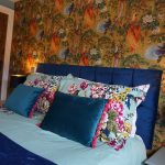 King-size bed with a blue velvet headboard, floral bedding, colourful bird wallpaper and tiger bedside lamps.